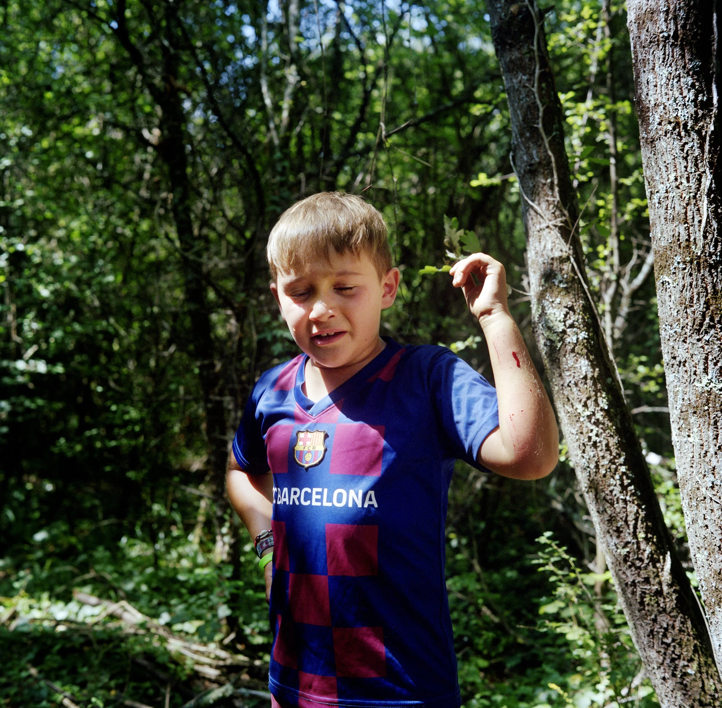 A happy-faced boy wearing an FC Barcelona soccer jersey, standing in a forest, eyes squinted and hand raised, probably injured or having left a difficult situation.
