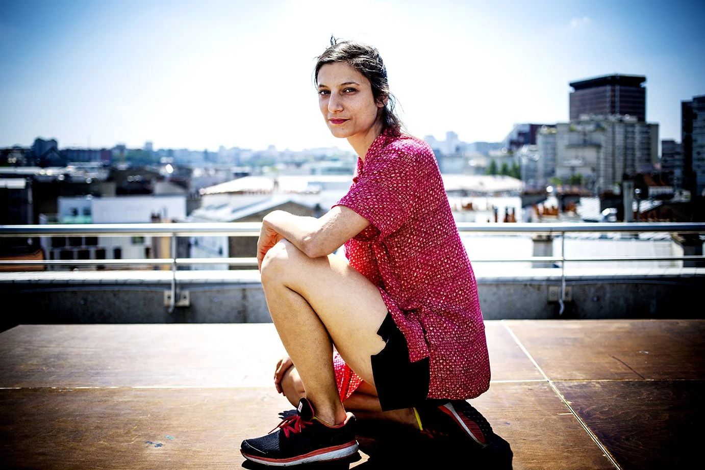 Young woman in a patterned red dress, sitting on a rooftop with an urban view in the background, wearing sports shoes. Sun and blue sky.