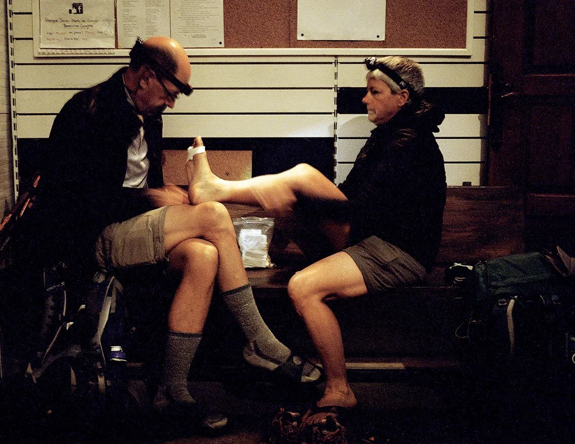 Two people sitting on a bench, one of them, a woman, holds her raised foot towards the other, a man, in an indoor environment. The scene seems unexpected or humorous.