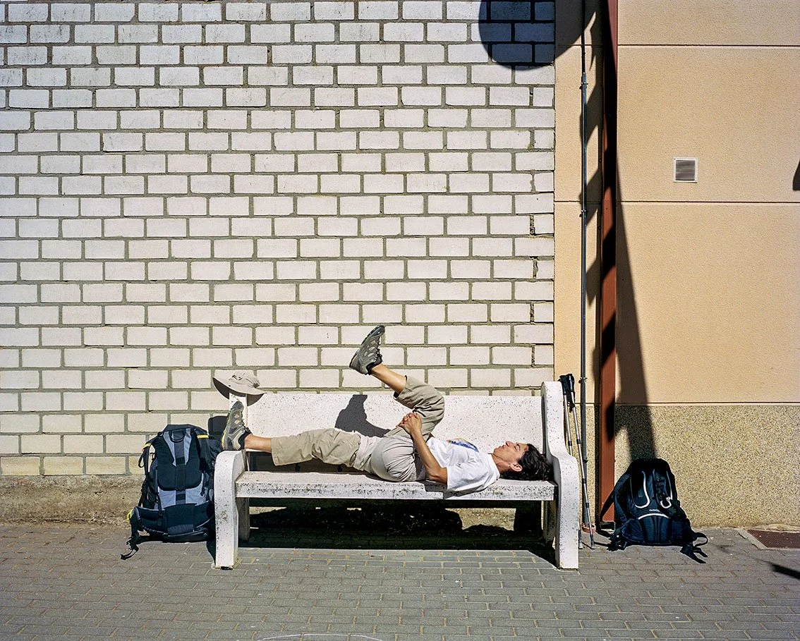 A man sitting and lying on a public bench, with a backpack beside him, resting.