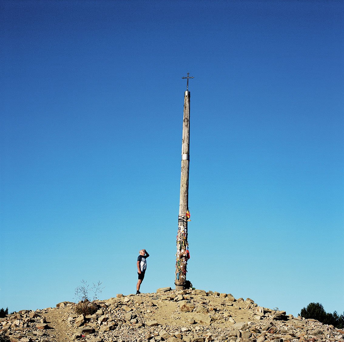 One person looks up while standing next to a wooden mast adorned with various charms and ribbons, on rocky ground, under a clear, blue sky.