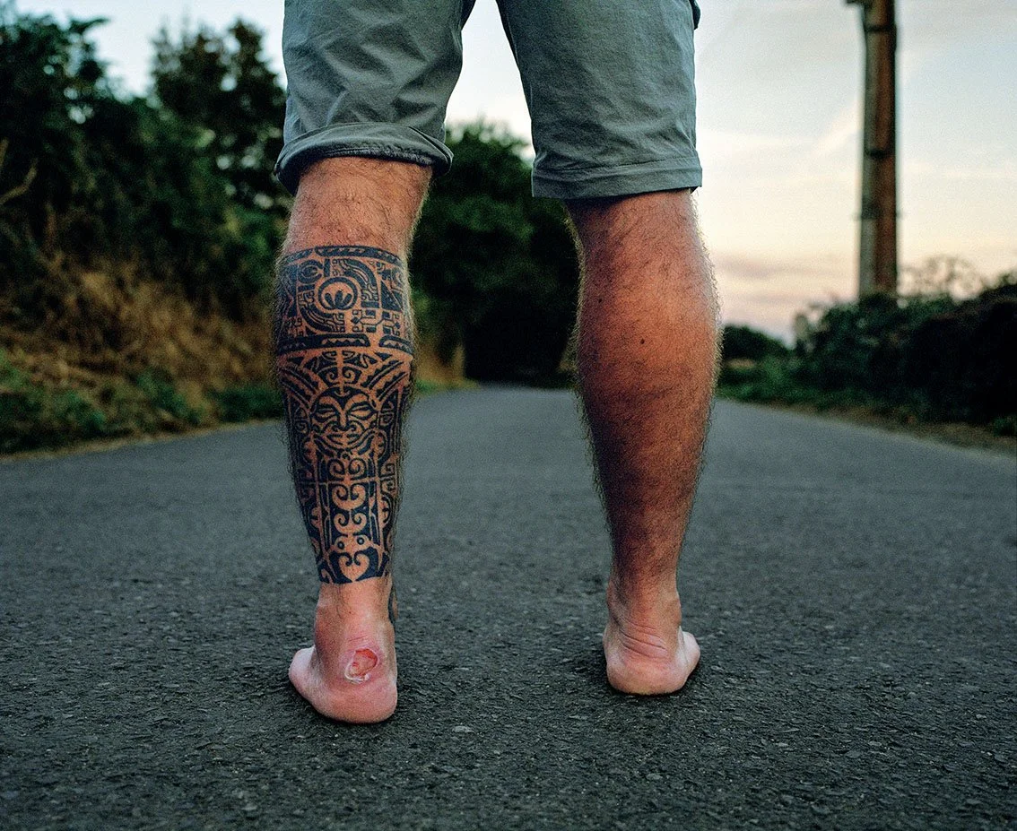 A man walks barefoot on a road, wearing gray shorts, with one leg half tattooed. The scene seems to take place at sunset, in a rural setting.