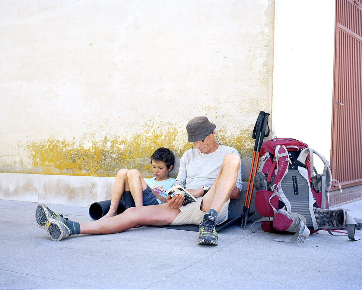 A man and a boy sitting on the sidewalk next to a backpack, reading and looking at a book under a tree, in an urban environment.