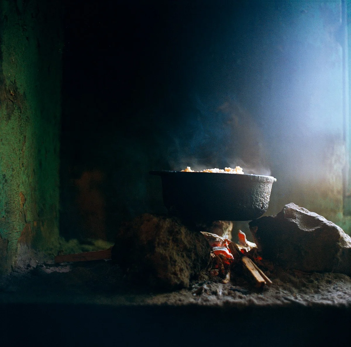 A black saucepan on a wood fire with glowing logs, in a rustic kitchen environment lit by natural light.