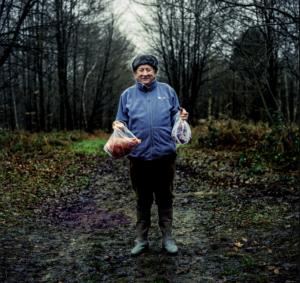 Un homme debout dans une forêt, tenant deux sacs en plastique remplis de viande, portant un pull bleu et des bottes.
