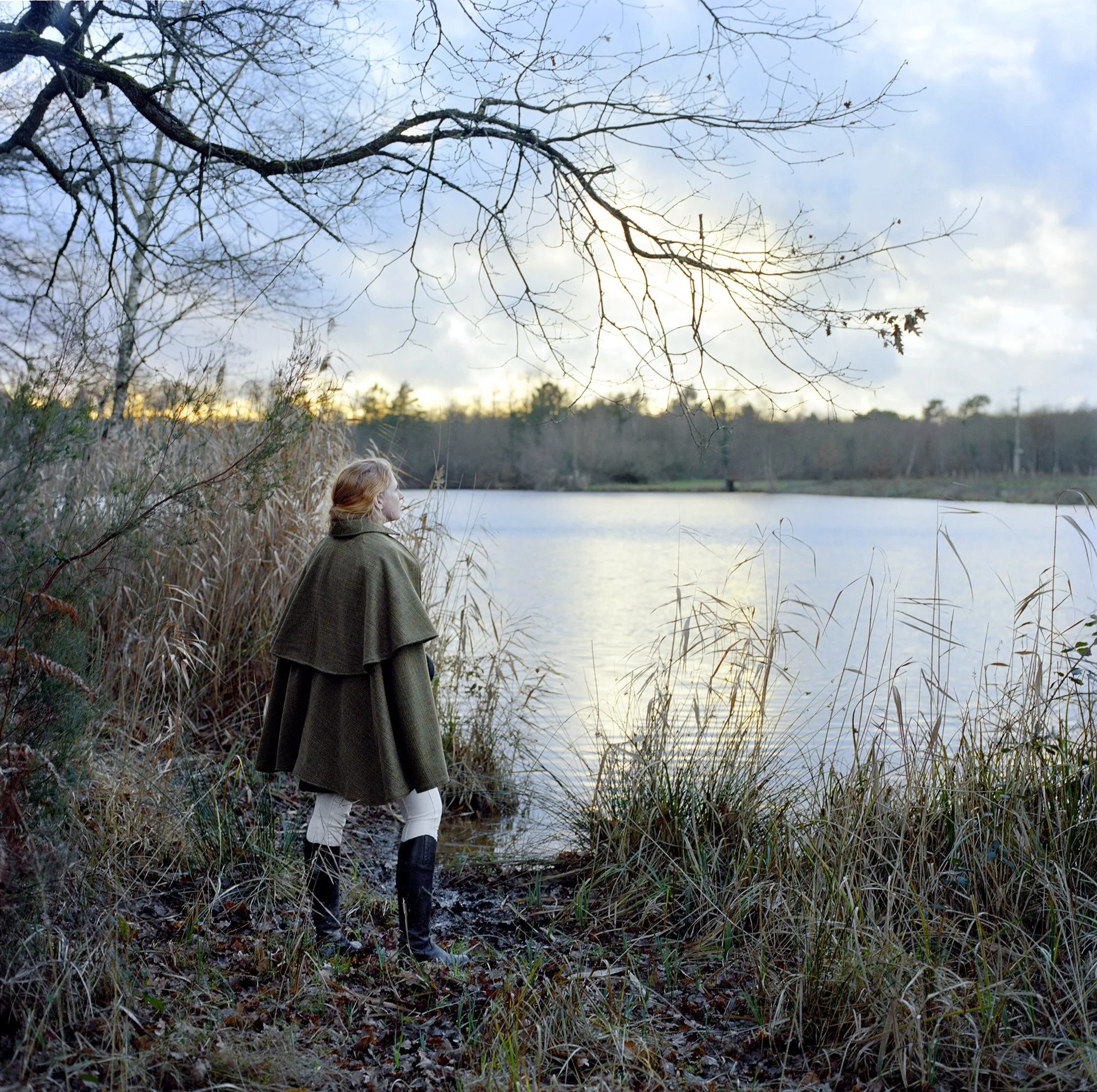 Une femme en vêtements d'hiver se tient au bord d'un lac, entourée de végétation sèche, observant la scène au crépuscule.