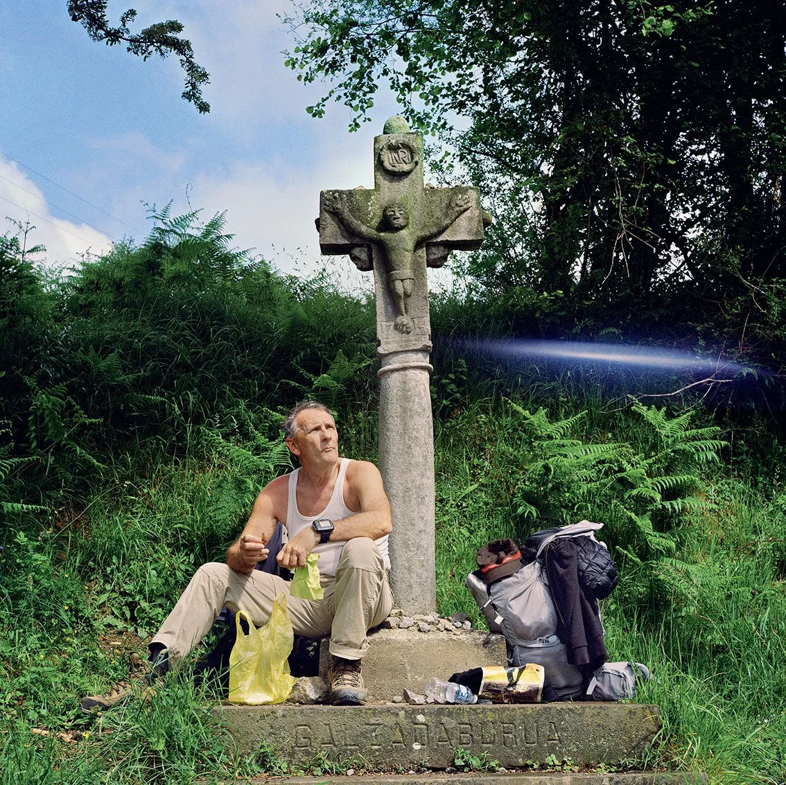 Man seated next to a stone cross with leaves and a backpack in a verdant natural setting