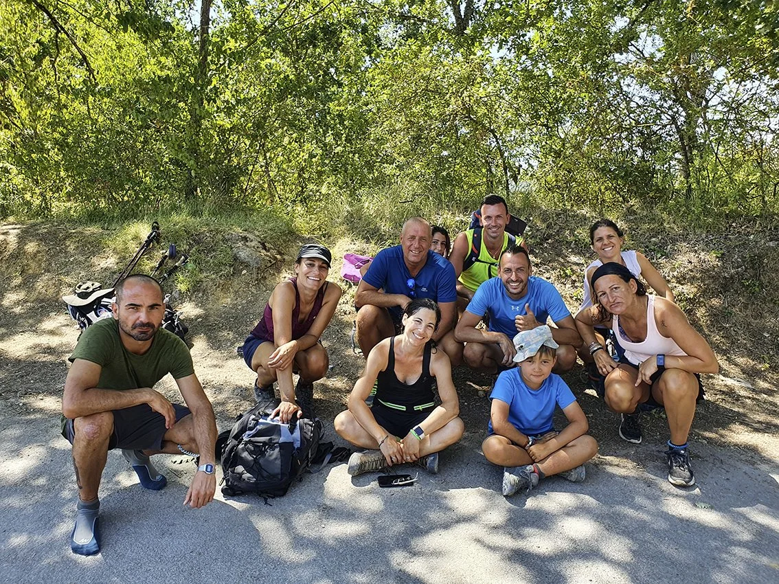 Group of smiling people posing outdoors under a tree during a hike, with backpacks and sportswear.