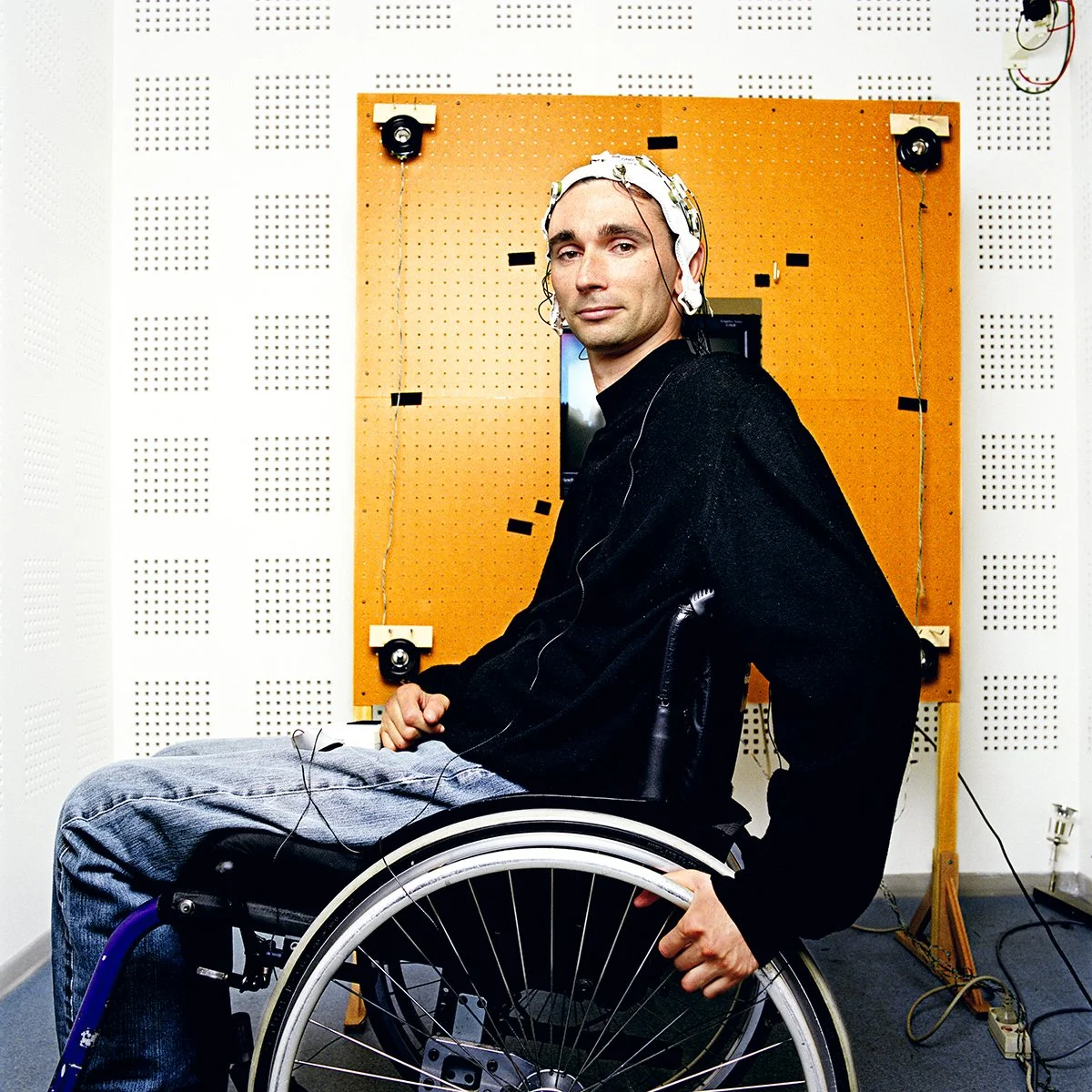 Man in wheelchair with electrodes on his head, likely participating in a brain-computer interface test in a laboratory.