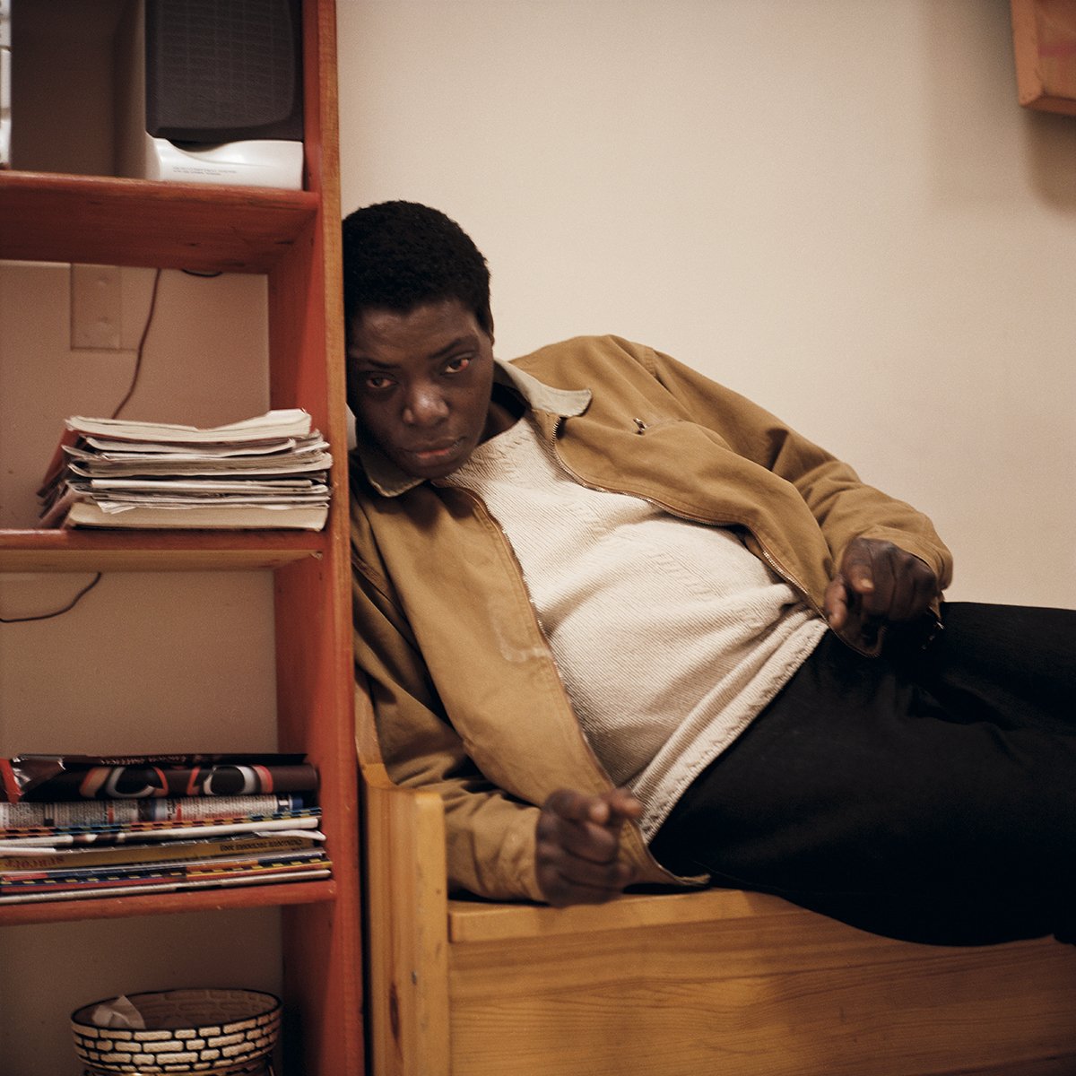 Un homme allongé sur un lit à côté d'une étagère en bois remplie de magazines et de papiers, regardant vers la caméra avec une expression sérieuse ou interrogative.