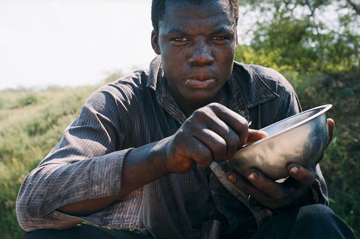 Young black man eating from a metal bowl in the open air, with a natural landscape in the background.