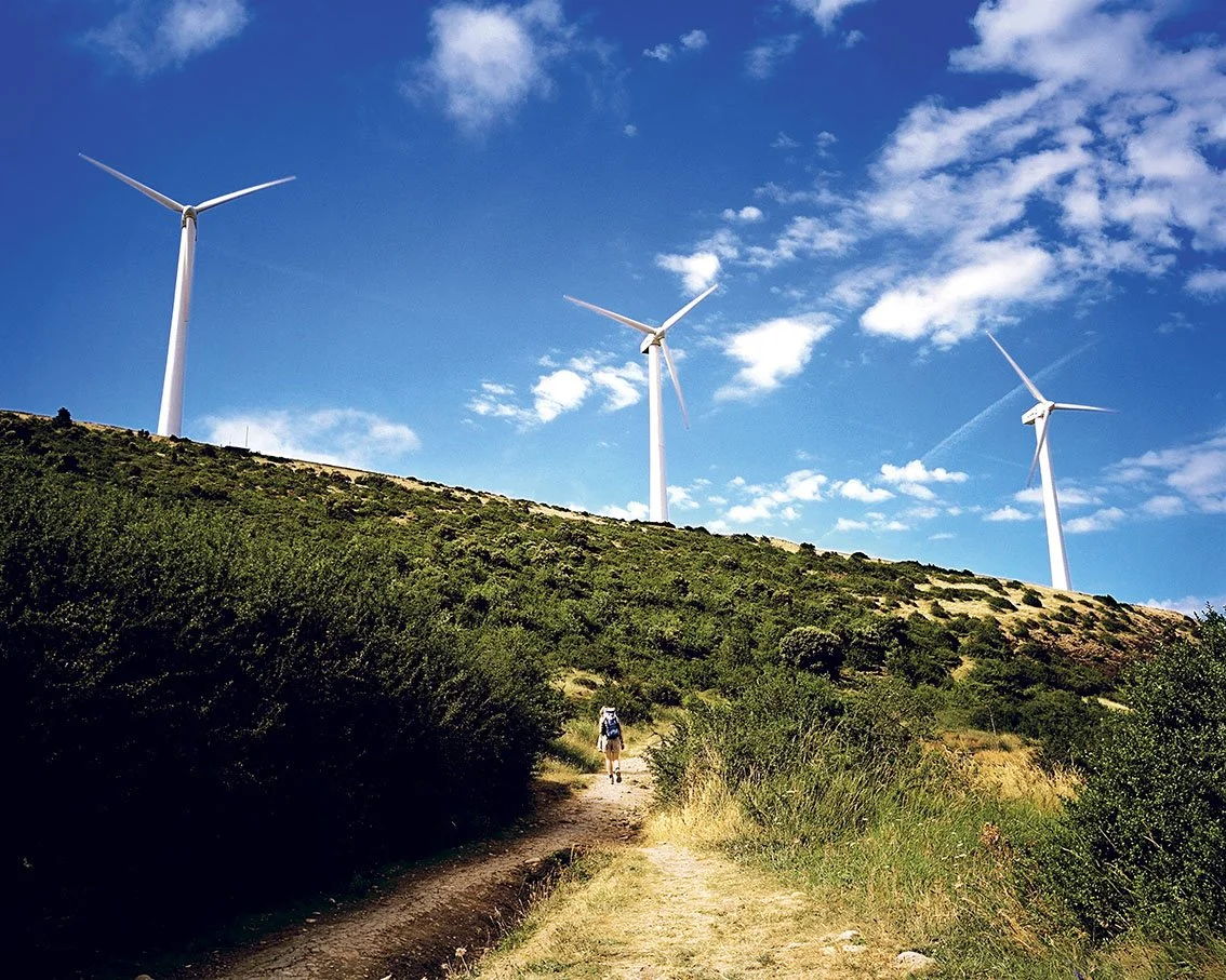 Dirt track with a walker carrying a backpack, descending a hill with wind turbines in the background, under a blue sky with a few clouds.