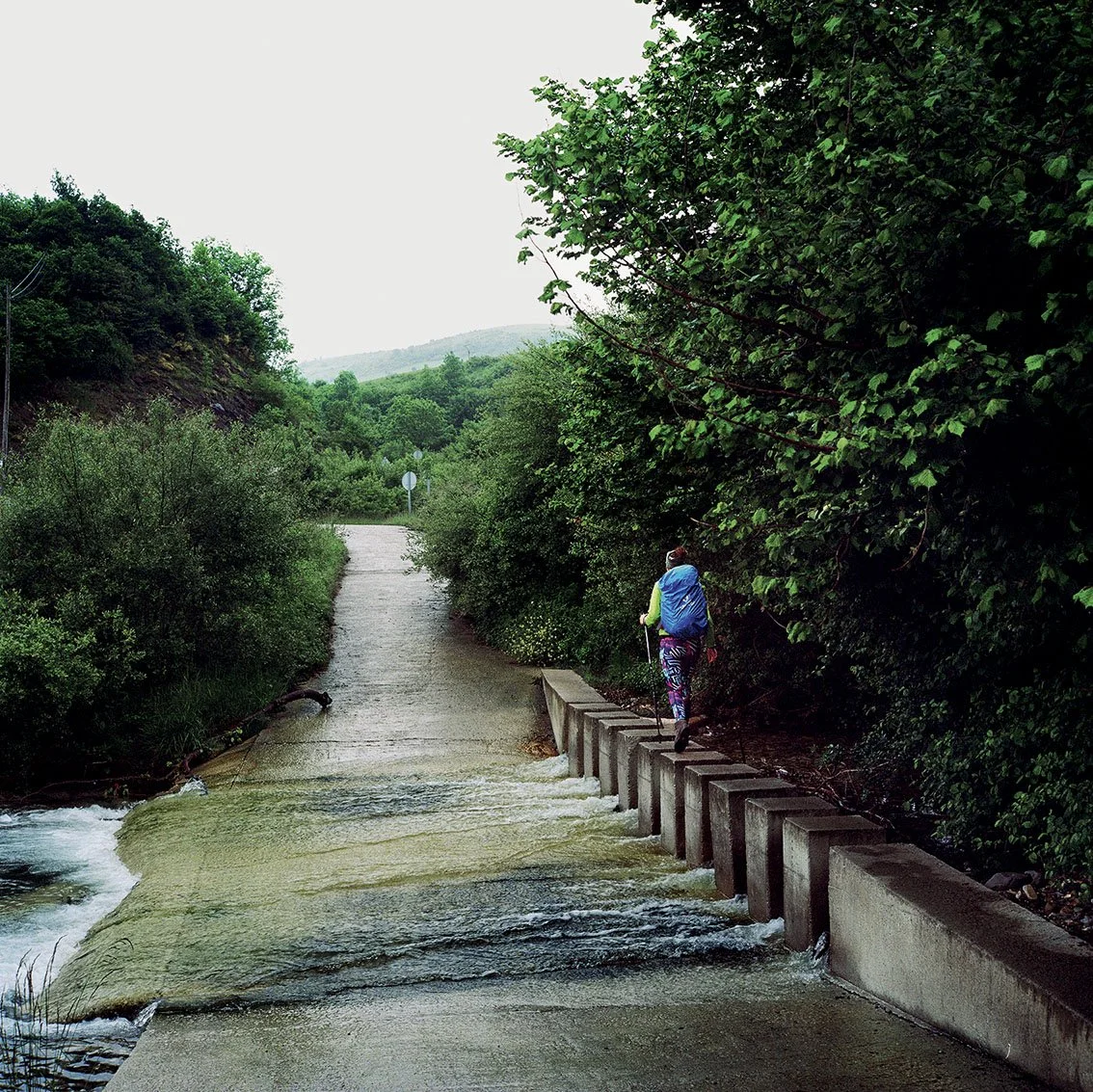 A person with a backpack walks along a path lined with trees and bushes, near a small waterfall or stream in lush natural surroundings.