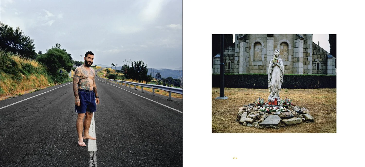 Homme tatoué en short debout sur une route vide, statue religieuse avec fleurs devant un bâtiment en pierre.