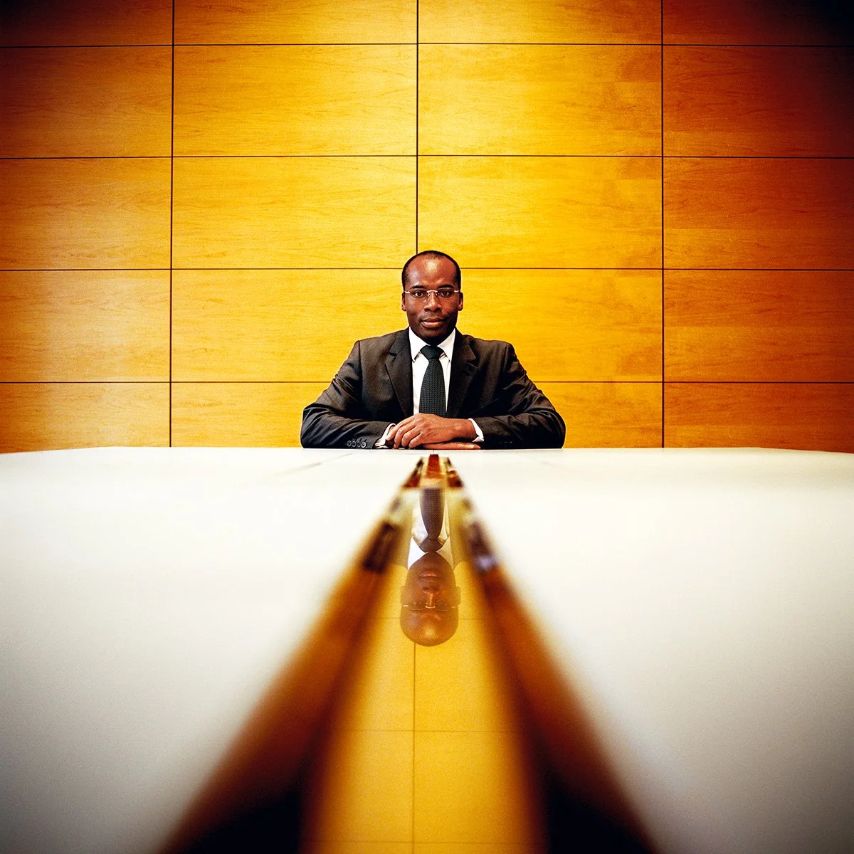 African businessman in black suit seated at a large table, with a wooden wall behind him, and his reflection visible on the table.