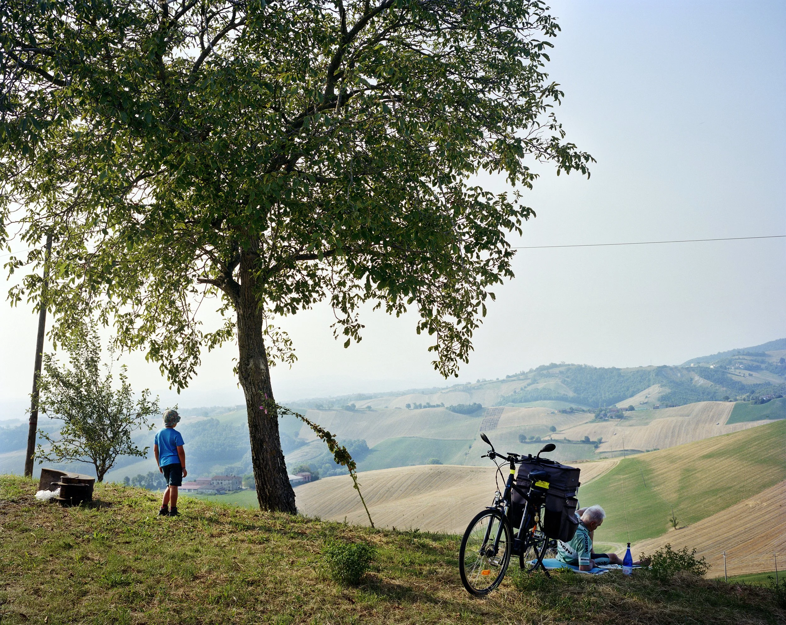 An elderly woman sits on a blanket next to her bicycle, drinking a bottle of water, with a child looking at the landscape in the background, in rolling countryside. A tree provides shade and cultivated fields stretch as far as the eye can see.