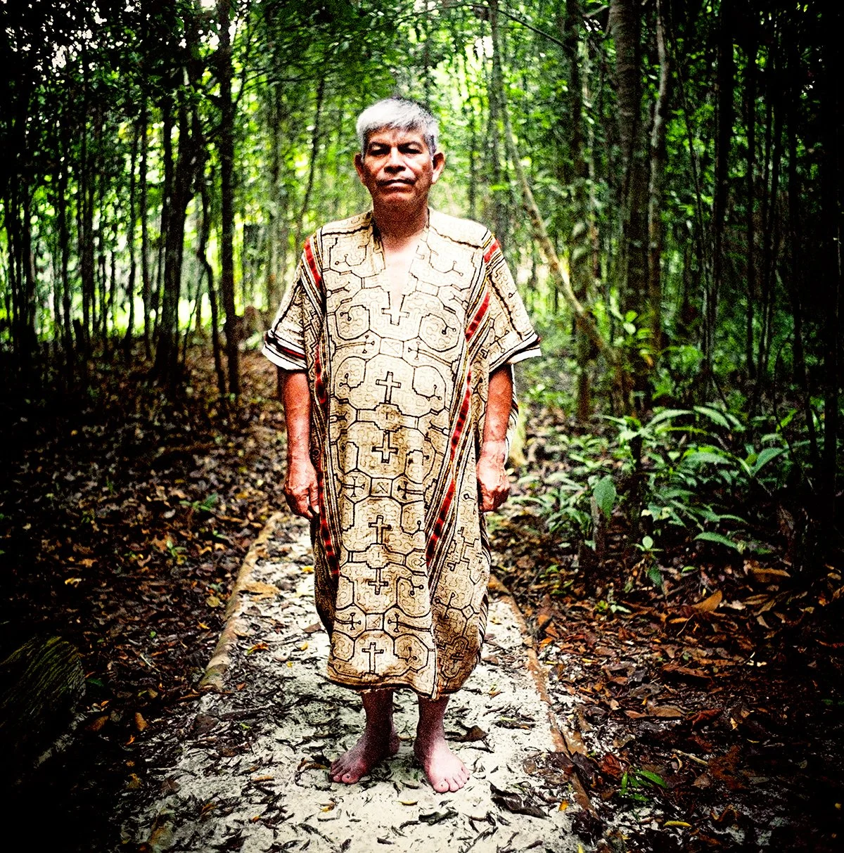 A native man standing on a forest path, wearing a traditional dress with geometric patterns, in a dense jungle.