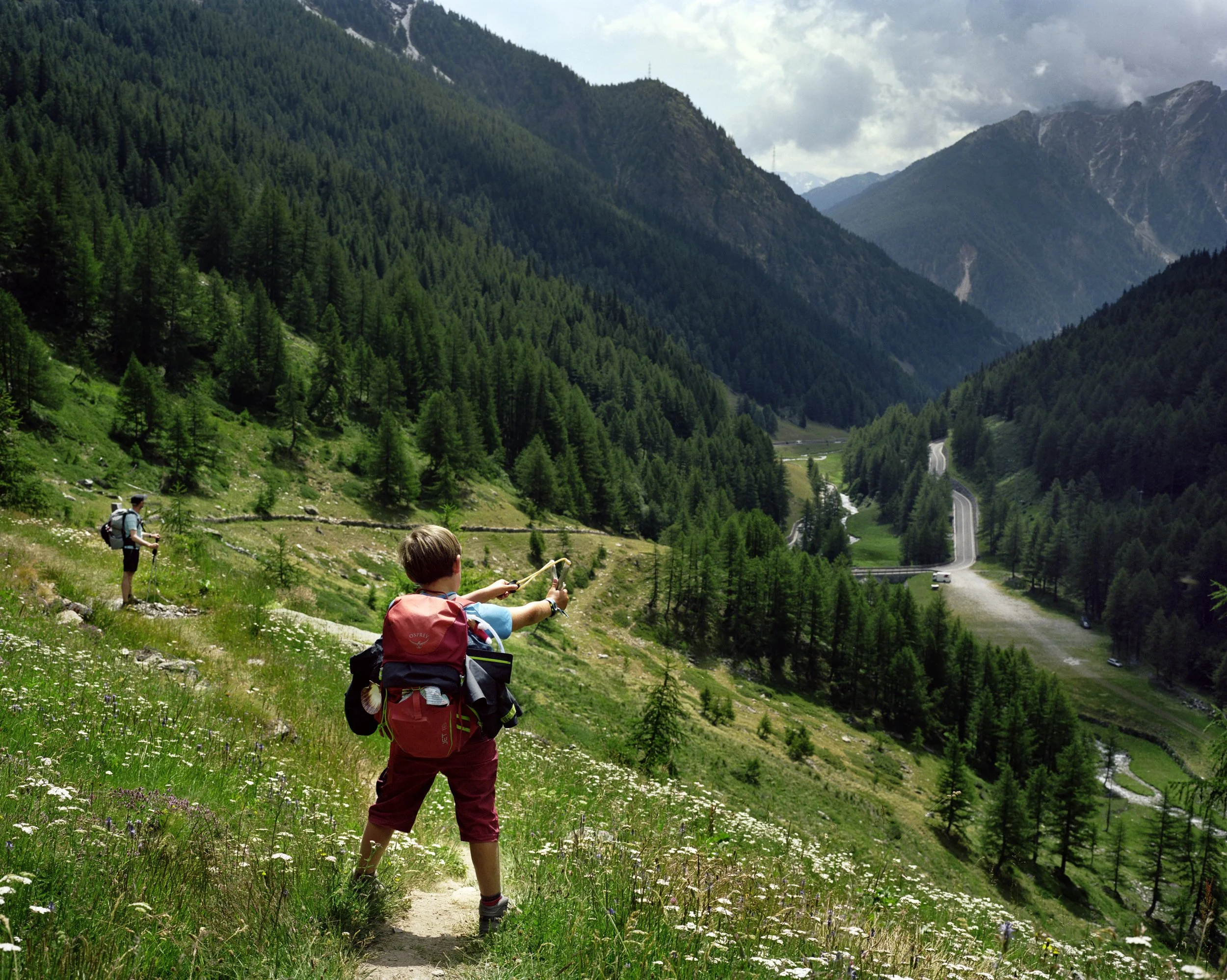 Two children with backpacks hiking in a green mountain valley.