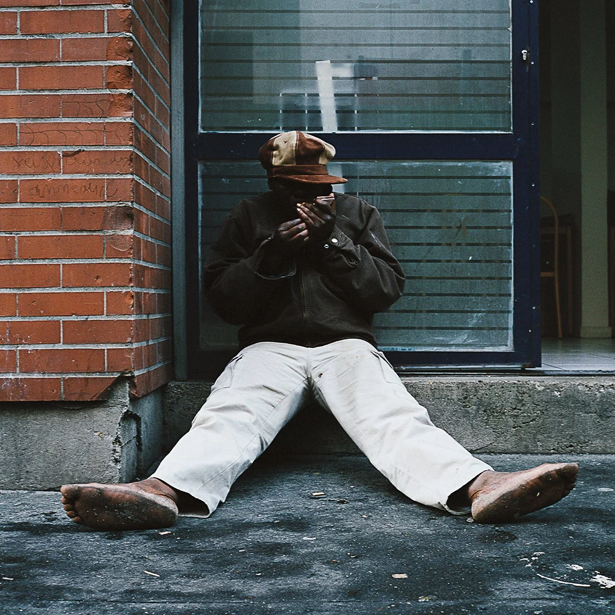 Une personne assise par terre contre un mur en briques, portant une veste sombre, un pantalon blanc et un chapeau beige et marron, regarde un téléphone portable.