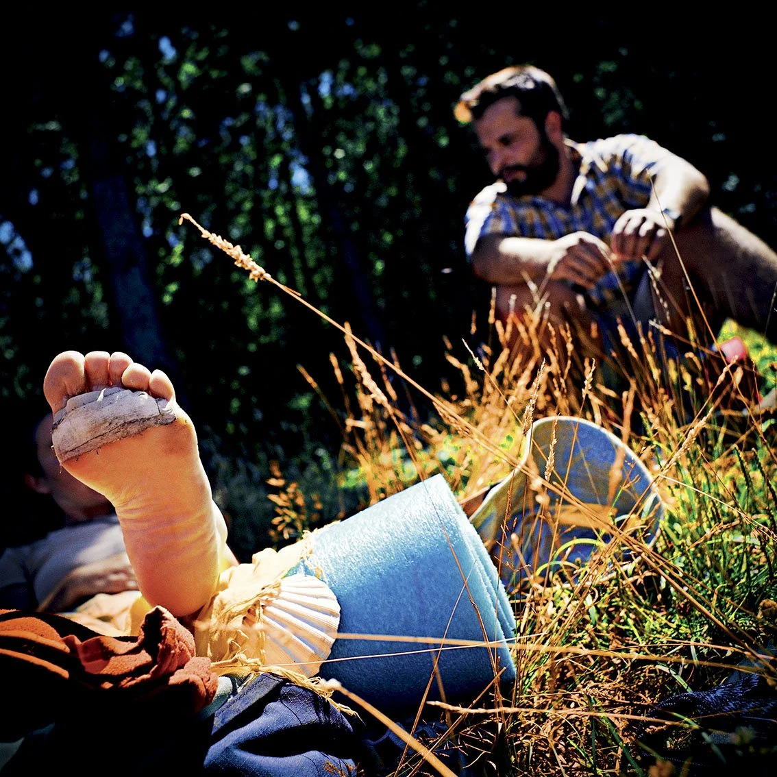 A man in a forest gathering mushrooms near a reclining person holding a mushroom in his hand.