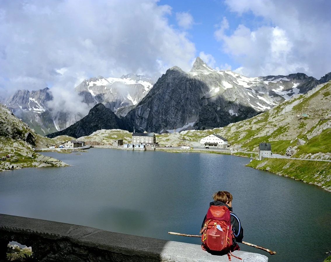 Young hiker with a red backpack, sitting on a wall looking out over a mountain lake surrounded by snow-capped peaks, in an alpine landscape.