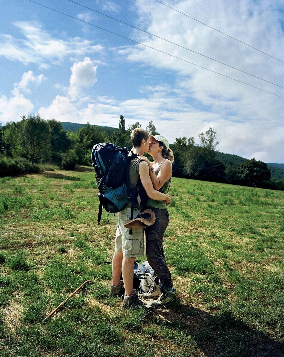A couple on a trip, kissing in an open-air field, with a blue sky and a few clouds, surrounded by nature and hills.