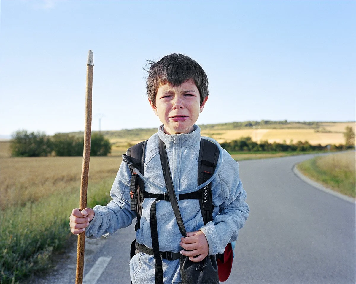 A boy with a backpack and a gray jacket, holding a long branch, crying on a country lane on a sunny day.