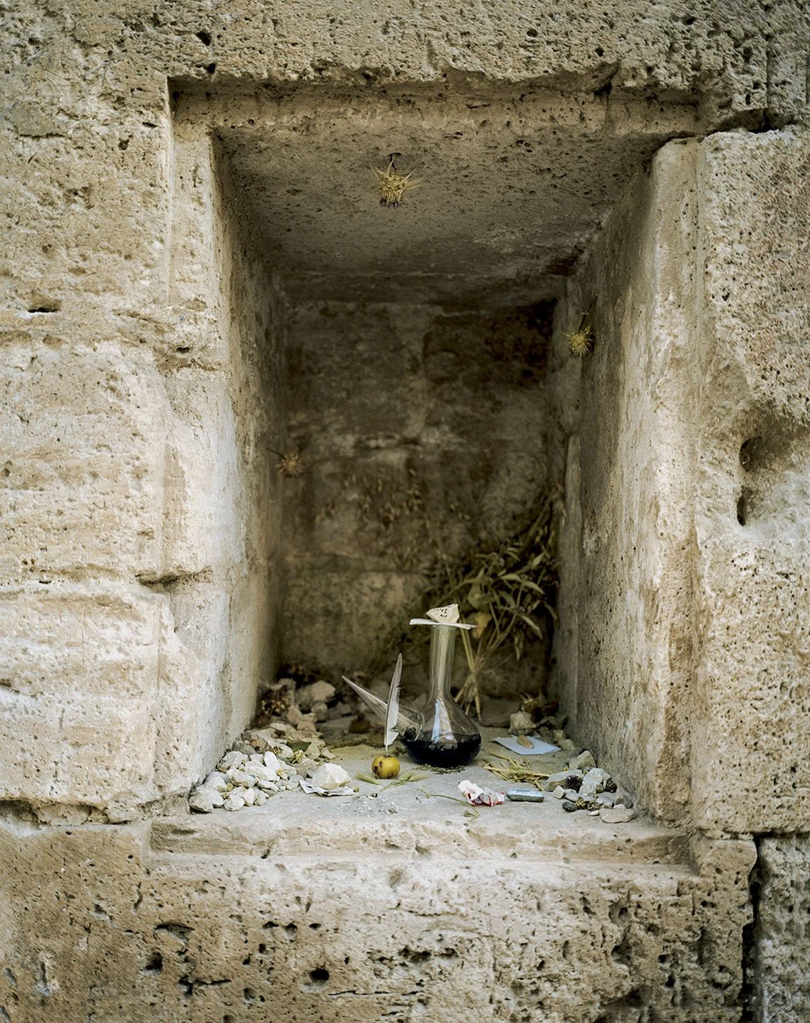 A niche in a stone wall containing a glass bottle, small objects and dry vegetation.