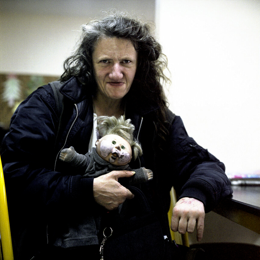 Une femme avec de longs cheveux bouclés tient une poupée aux traits hybrides humains et animaux. Elle porte une veste foncée et est assise à une table.