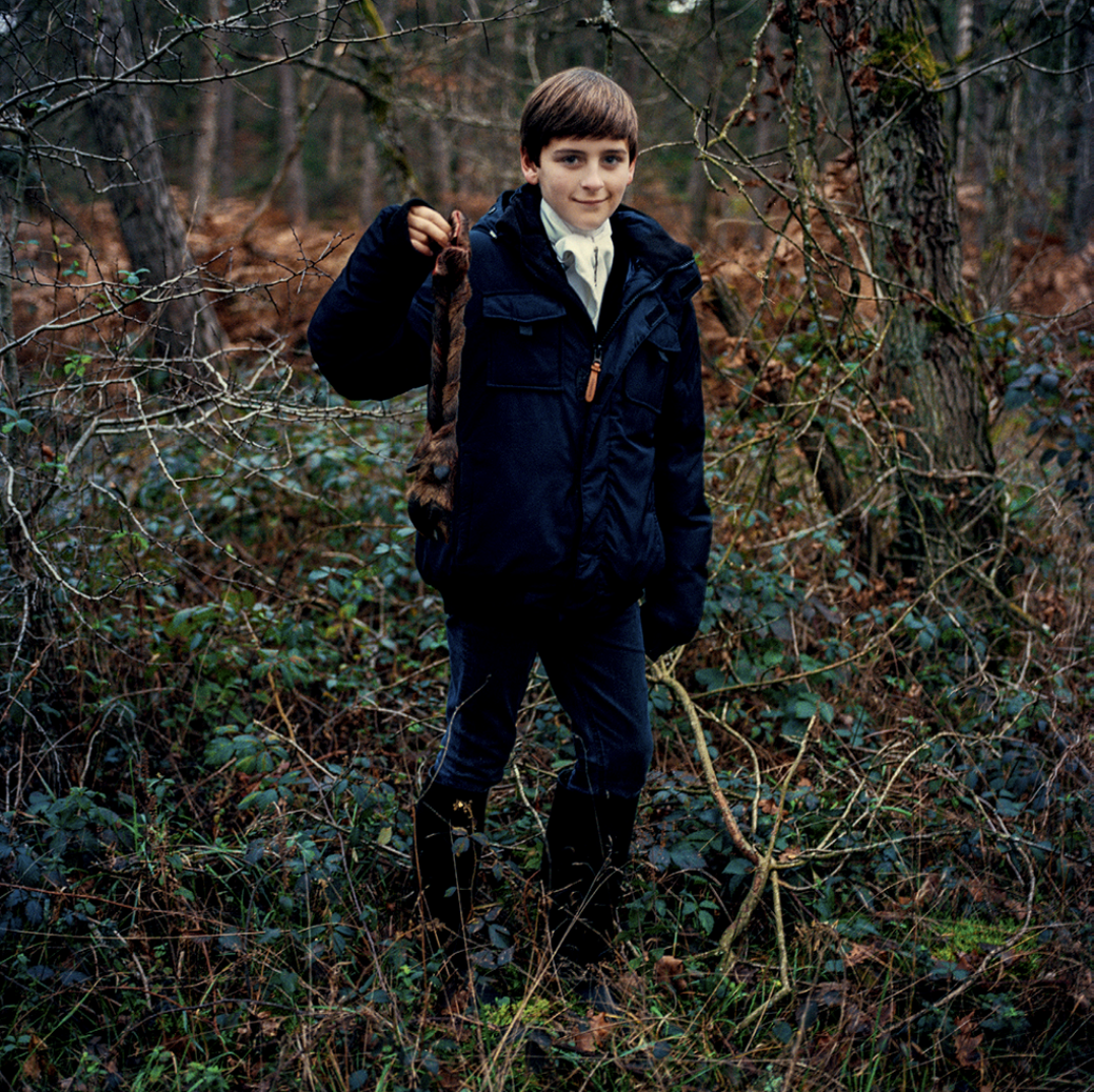 Jeune garçon dans une forêt, tenant un petit animal dans sa main, portant une veste noire, des pantalons foncés et des bottes, entouré d'arbres et de végétation dense.