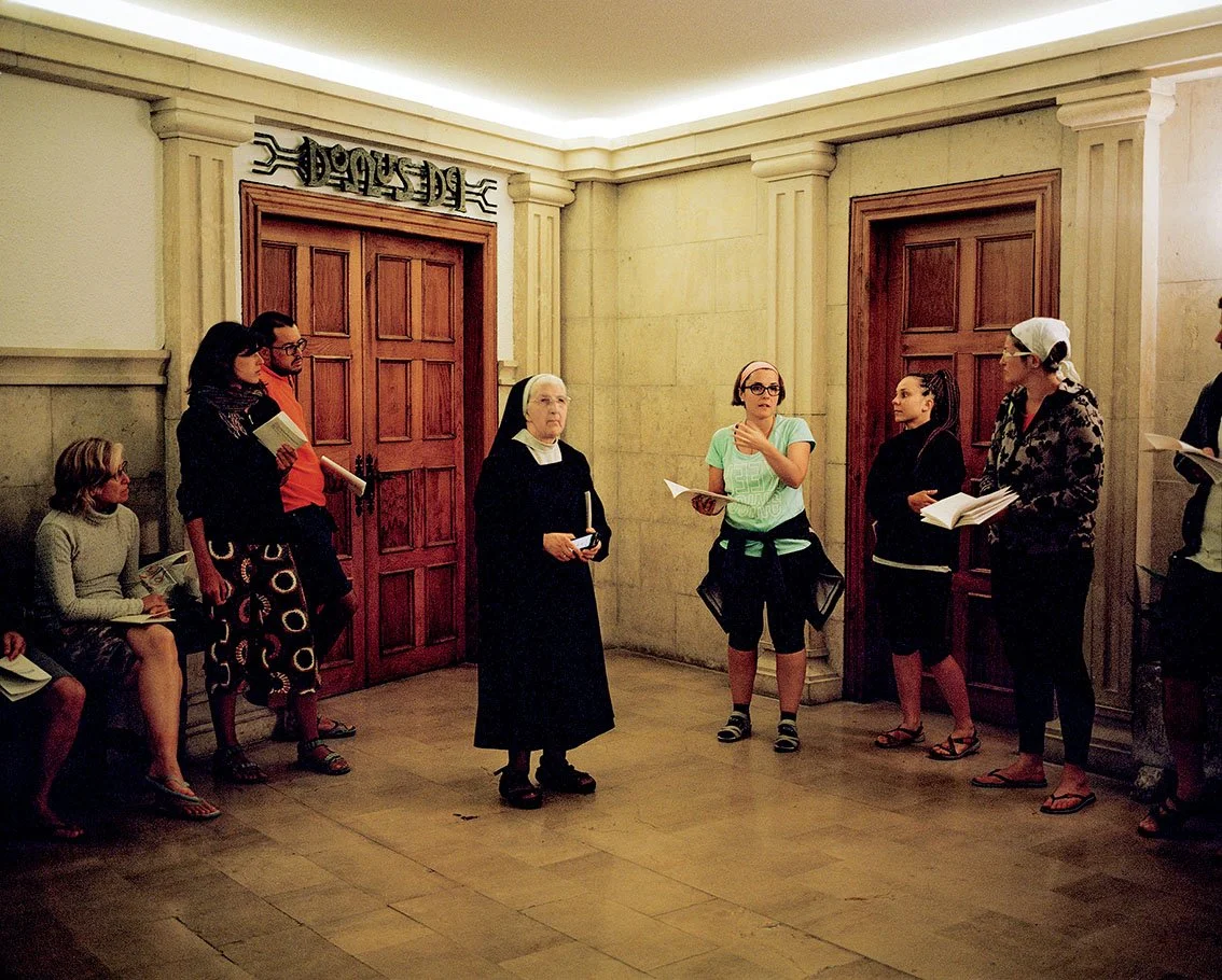 A group of people, including a nun, appear to be taking part in a meeting or discussion in a room with wooden doors. Among them are several women, a man and a young girl holding books or documents.