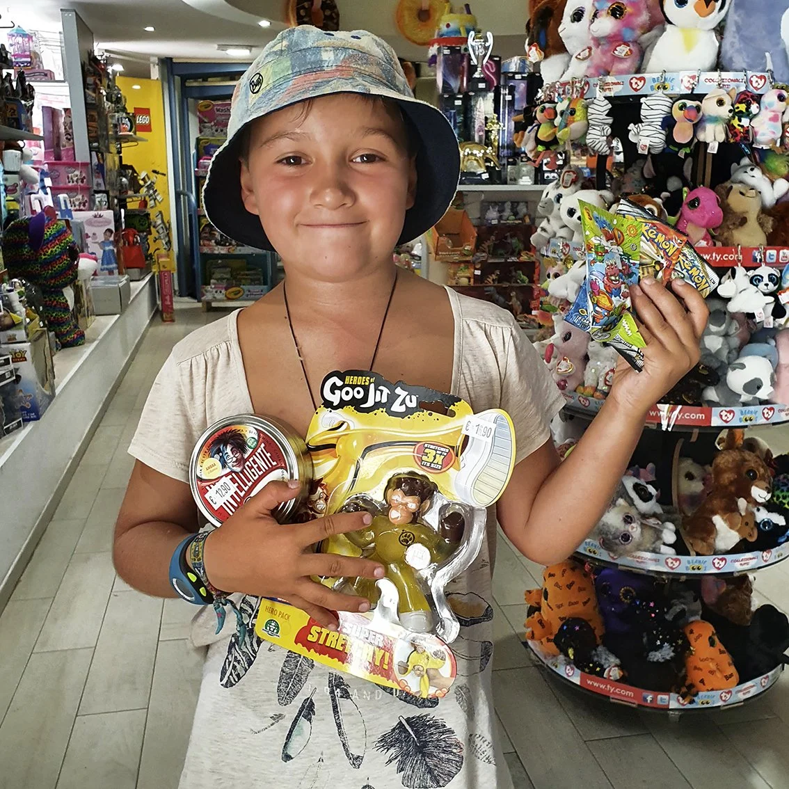 A smiling boy wearing a colorful hat, holding figurine toys and bags of candy in a toy store full of plush toys and children's items.