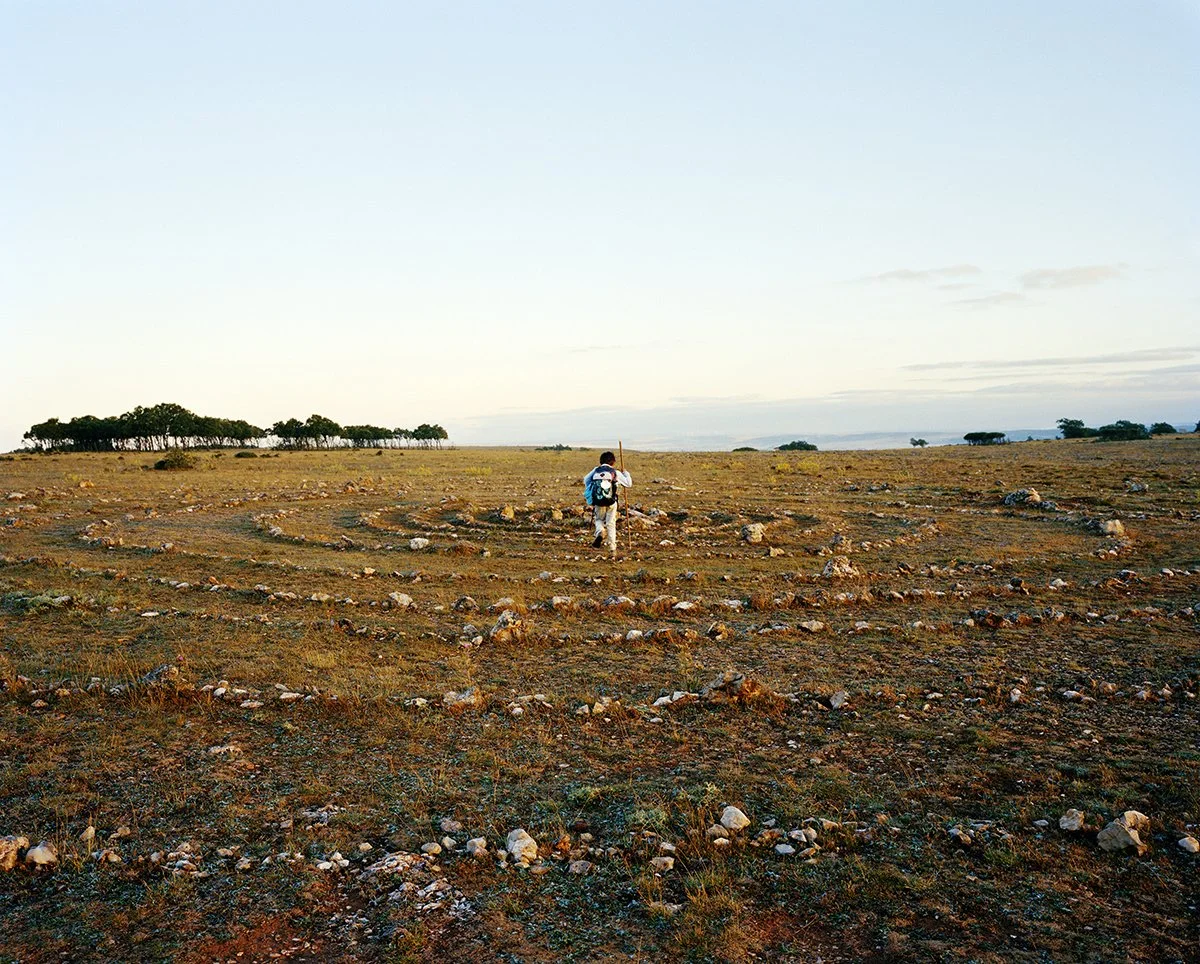A person walking alone in a vast desert or arid landscape with a backpack, holding a stick, under a clear evening sky.