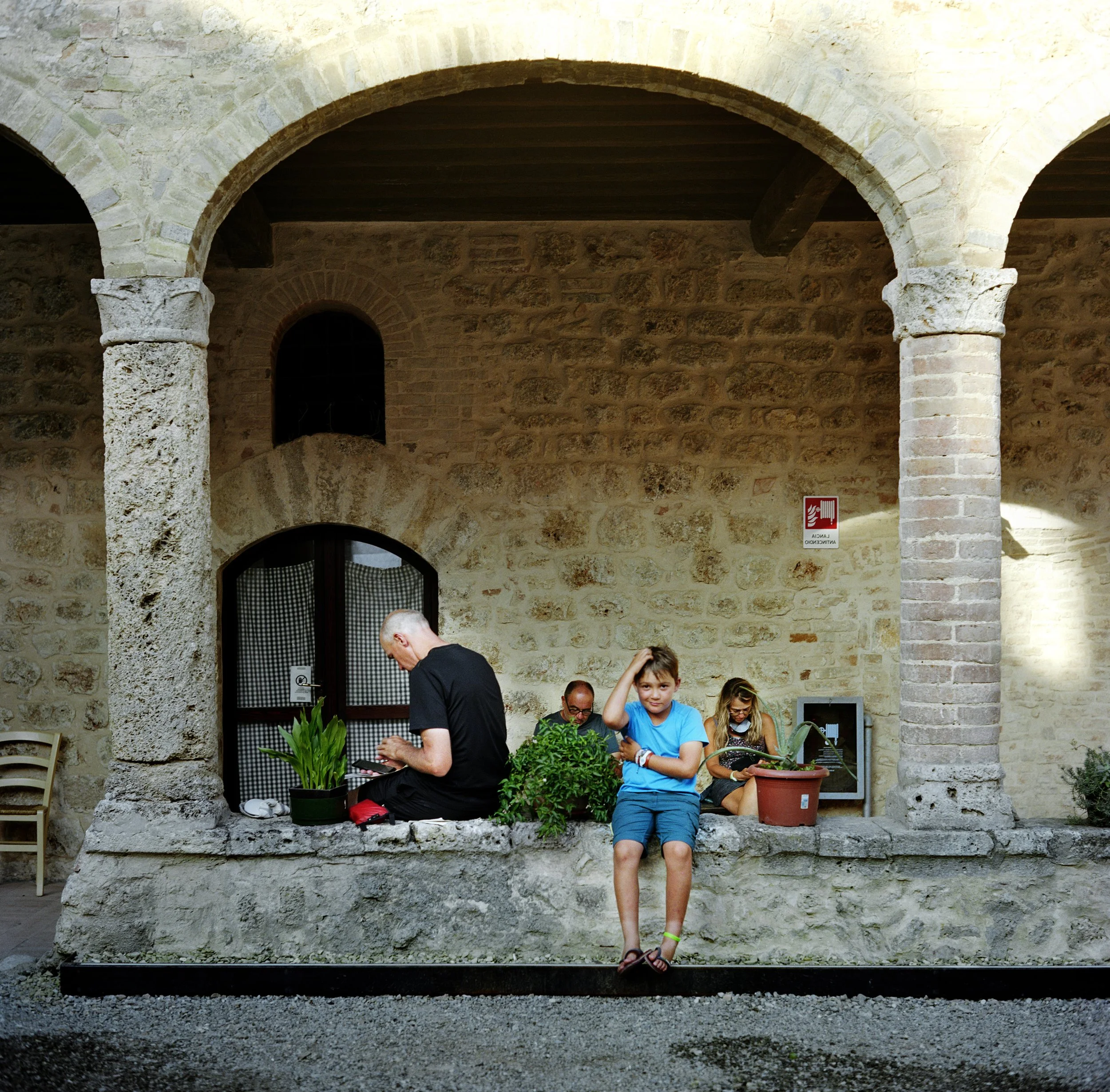 Four people sit on a stone structure under a stone archway, one woman and three men. A boy in blue sits at the edge, looking at the camera, a woman in a checked shirt and mask, a man in glasses and a woman e