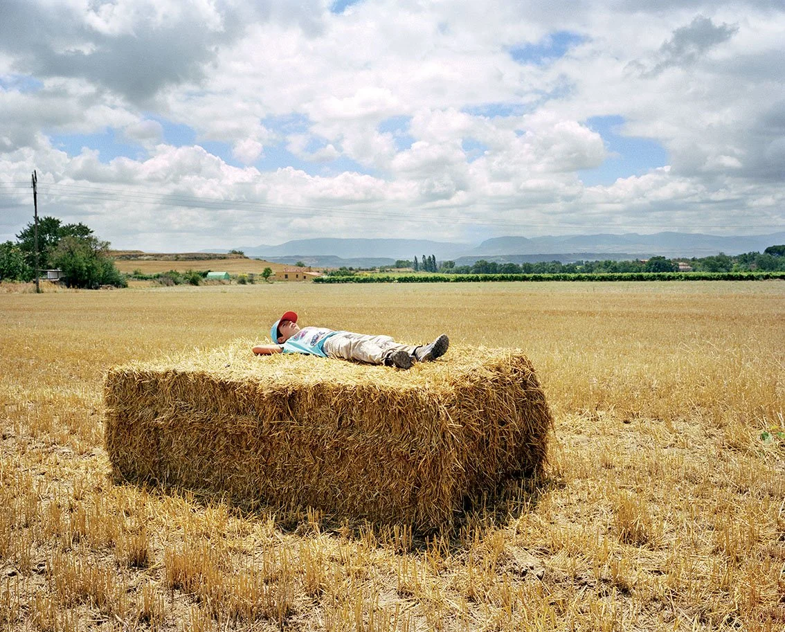A child lies on a large haystack in a field in broad daylight, with a partly cloudy sky and mountain horizons in the background.