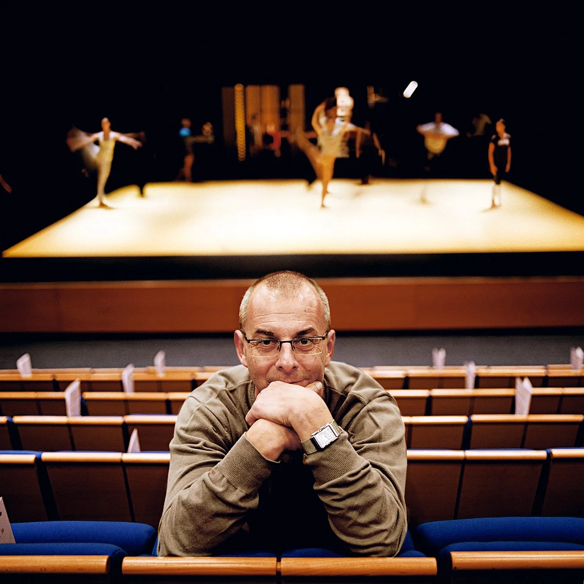A man sitting in a theater in front of the stage where a dance performance is in progress, front view.
