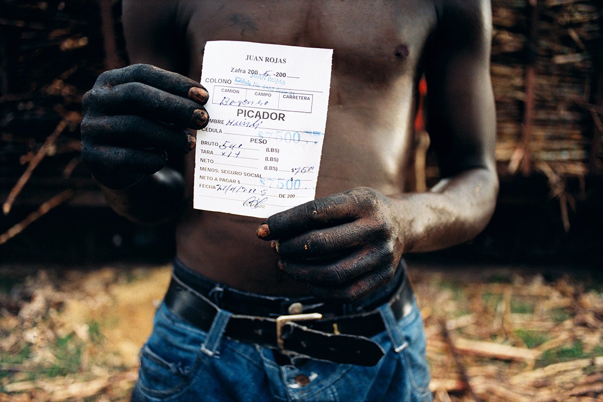 A naked man with very dirty arms and hands holds a sheet of paper or a written note, in an open-air environment.