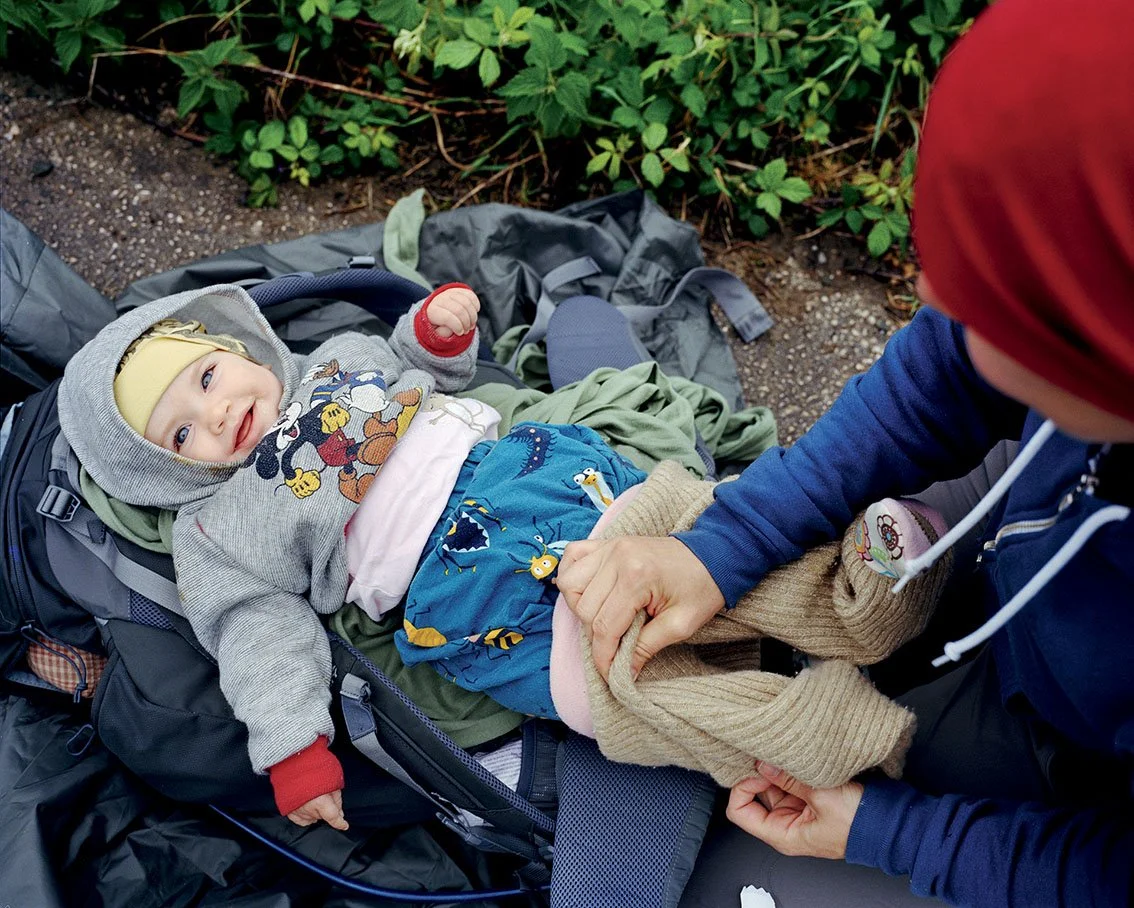 A baby lying in a backpack, dressed in a gray Mickey Mouse hoodie, yellow beanie, and blue pants with toy motifs, smiling and looking straight ahead. One person, probably an adult, is holding him by the belly, carrying a vee.
