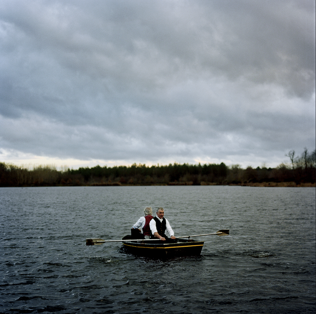 Deux personnes en costume traditionnel naviguent en bateau sur un lac, sous un ciel nuageux.