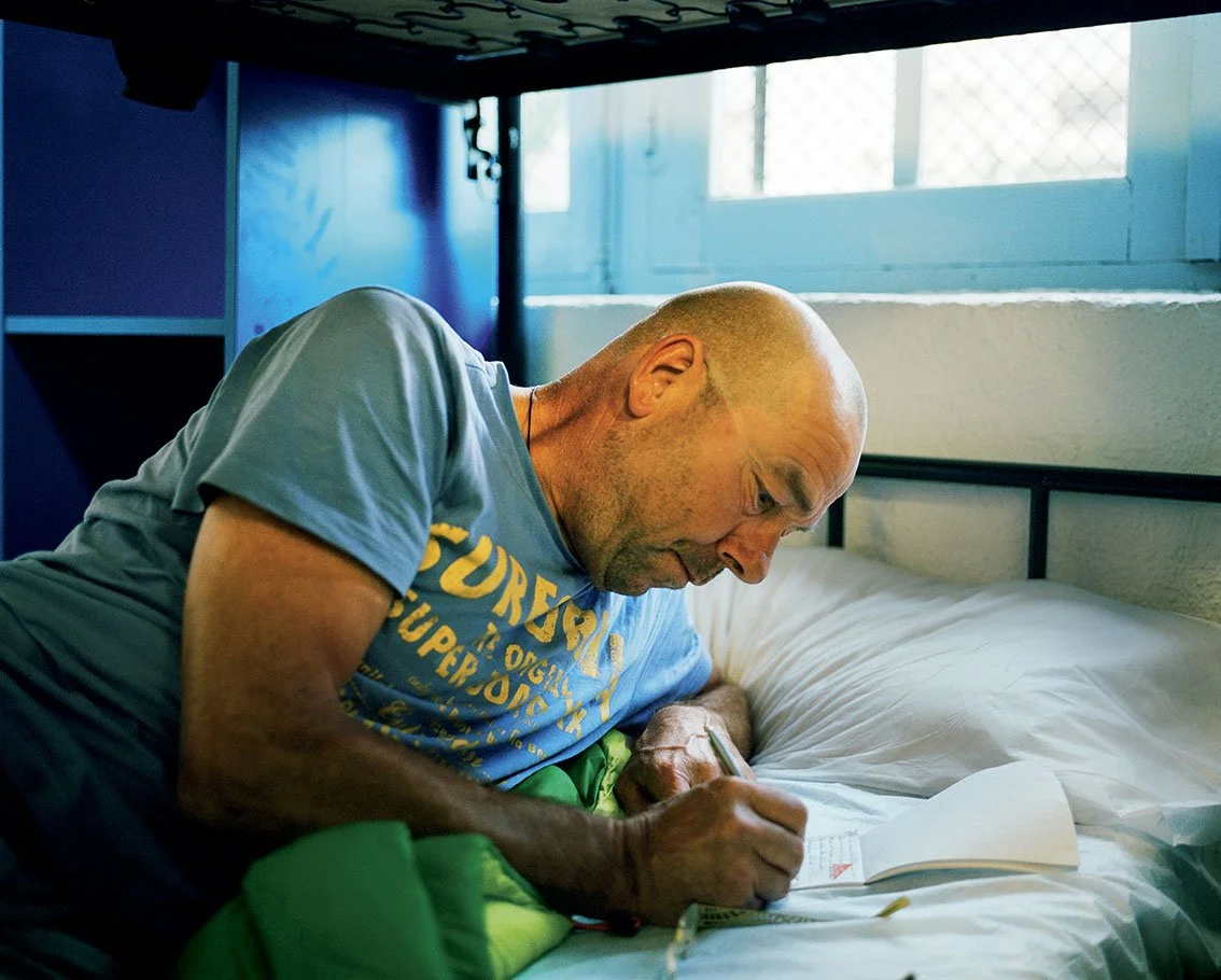 A man in a blue T-shirt writes in a notebook on a bed in a room with a window.