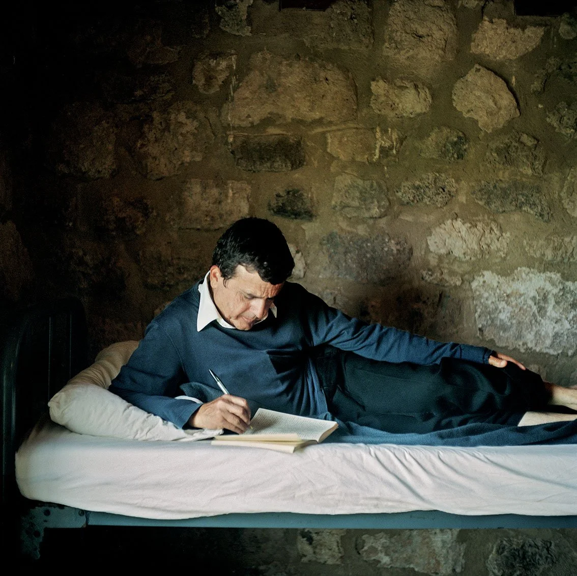 Man reading and writing in a newspaper while lying on a mattress in a room with a stone wall