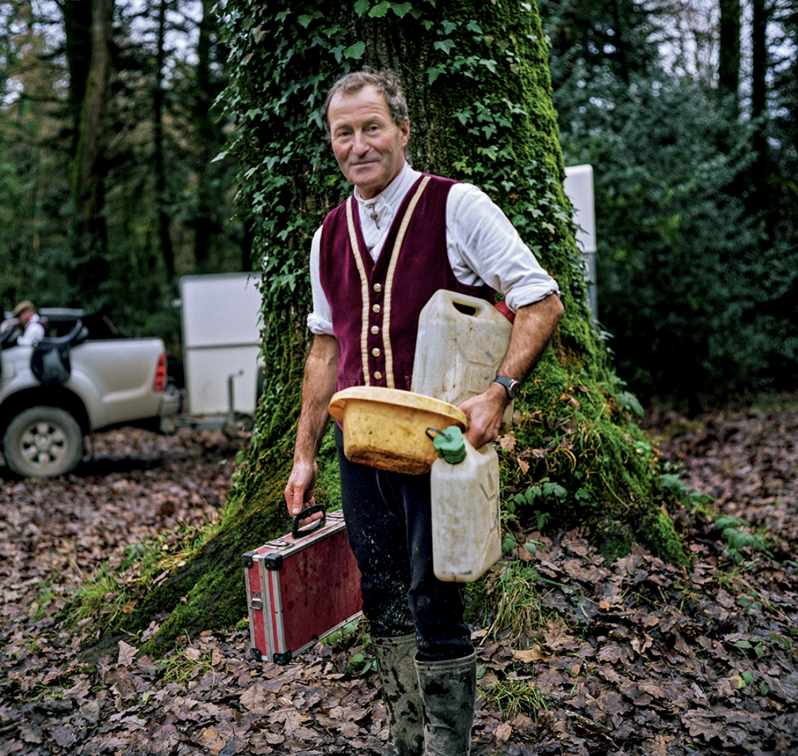 Un homme se tenant devant un arbre dans une forêt, portant un caddy, un récipient d'eau, une boîte à outils rouge, et vêtement de chasse ou de travail, avec une voiture en arrière-plan.