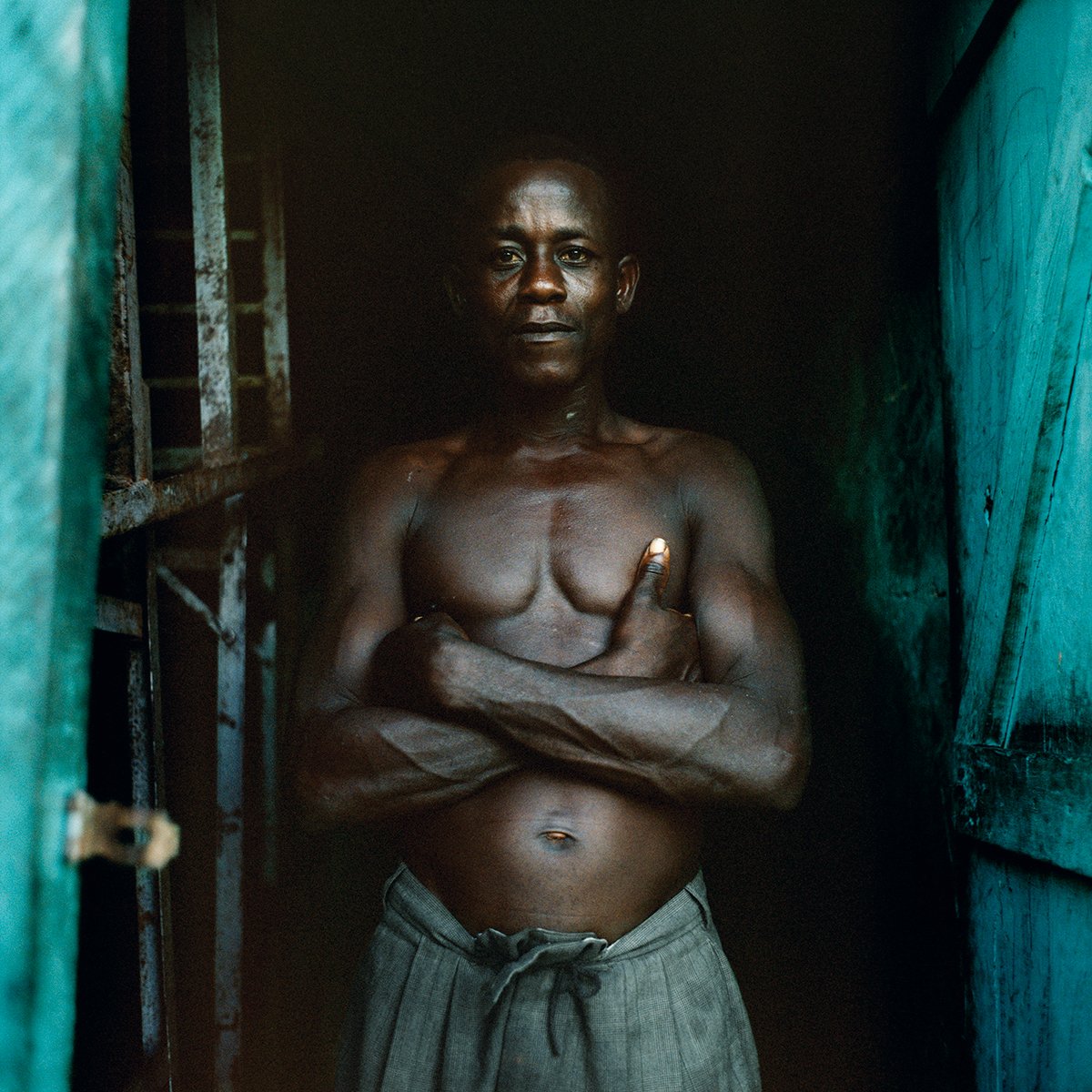 A naked man with folded arms stands in a small, dark opening, surrounded by wooden walls painted turquoise green.