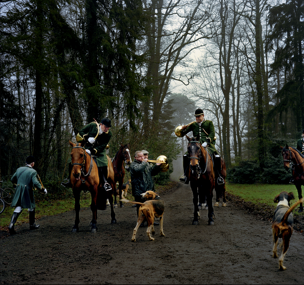 Groupe de personnes avec des chevaux et des chiens dans une forêt, quelques musiciens jouent de la trompette, ambiance brumeuse.