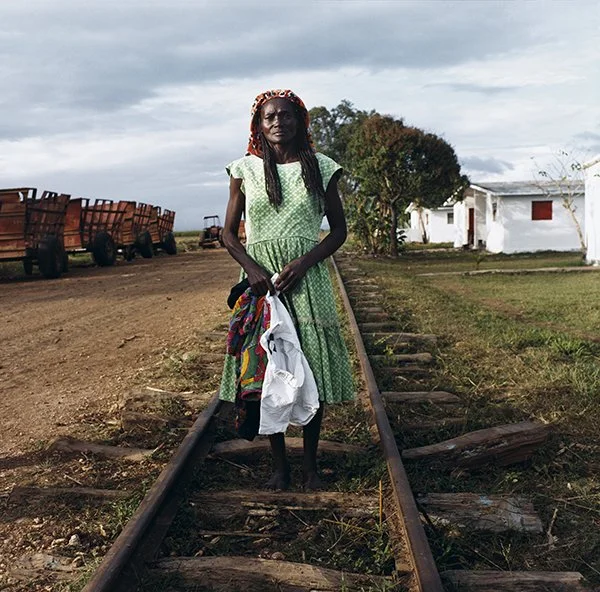 Femme âgée en robe verte, tenant des vêtements, debout sur une voie ferrée dans un paysage rural avec des maisons blanches et des wagons de train.
