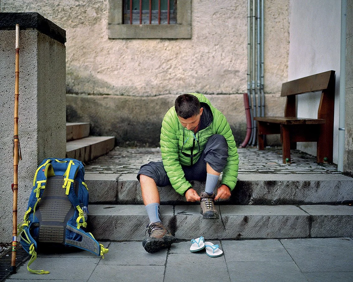Young man sitting on steps, putting on his shoes. Next to him, a pair of white sandals, a blue backpack with yellow details, a wooden walking stick, and a wooden skip.