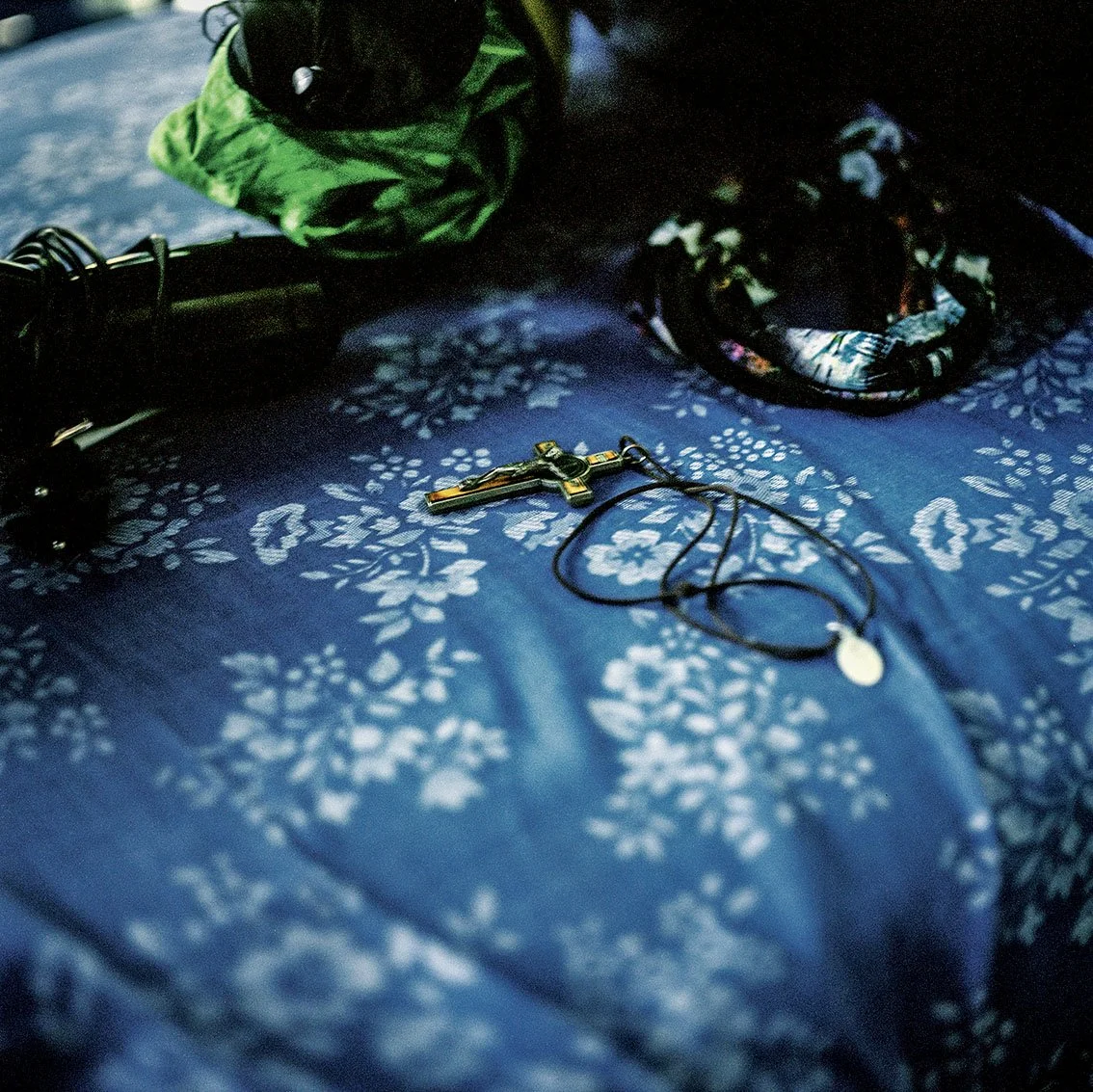 A cross and medallion on a blue cover with a floral motif, two hands partly visible in the background with green, patterned garments.