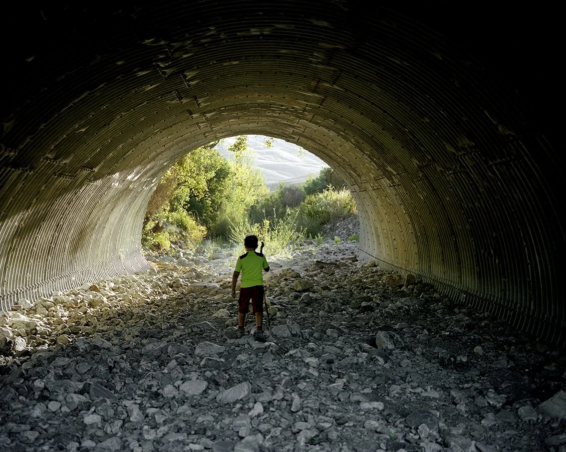 A child walks through a metal tunnel with a walking stick, leading to rocky terrain and abundant vegetation under a sunny sky.