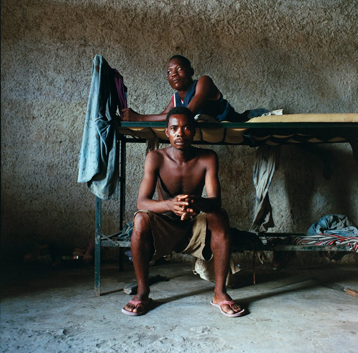 Two men in a modest bedroom, one seated on the floor, the other lying on a high bed, in a room with bare stone walls.