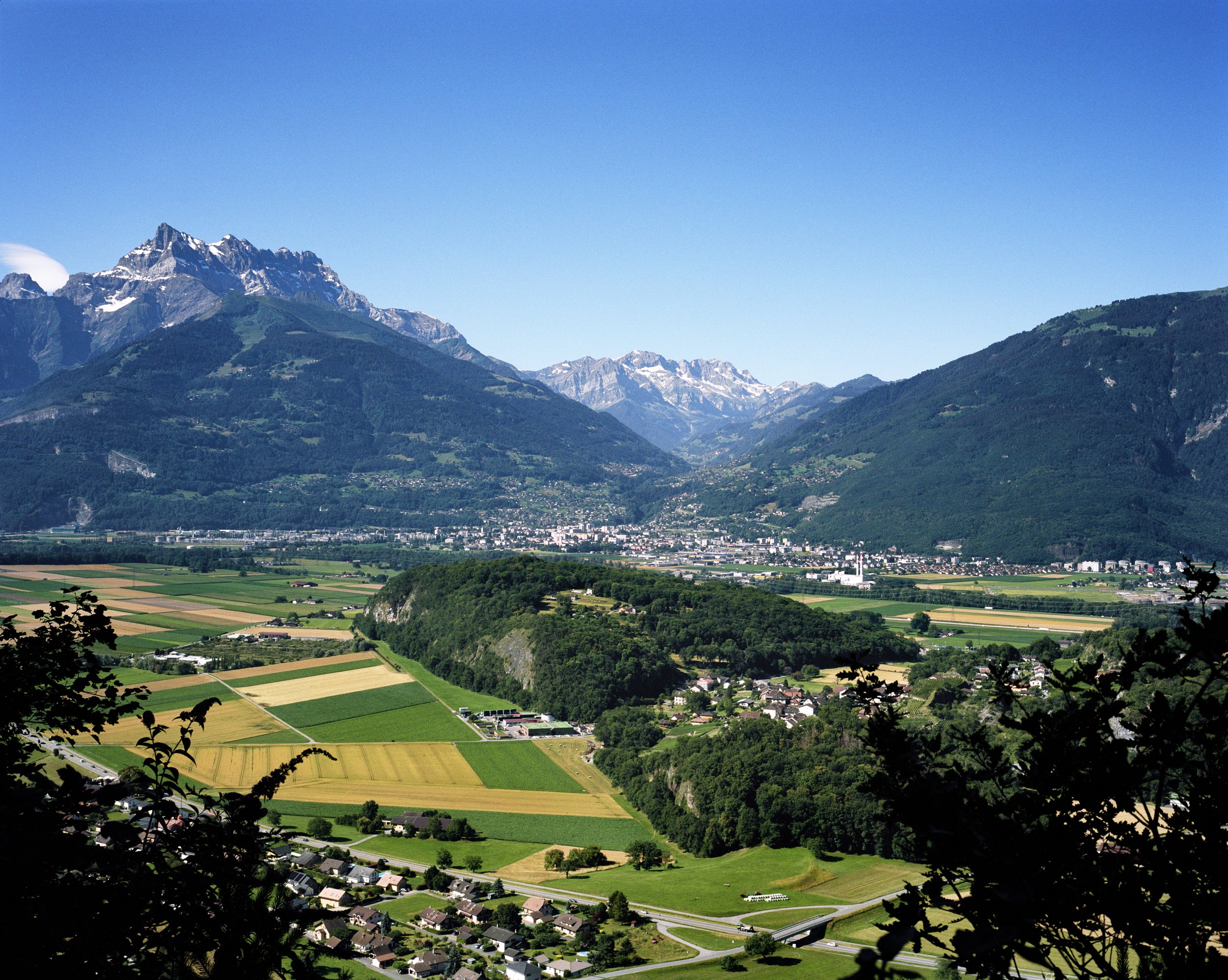 Mountain landscape with agricultural fields and a village in the foreground, light blue sky.